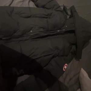 Svart dunjacka från Canada Goose XS - Säljer en svart dunjacka från Canada Goose i storlek XS. Jackan har huva, klassisk logga på ärmen och quiltad design. Perfekt för kalla dagar med sin tjocka och fluffiga känsla. Fodret är slätt och jackan stängs med dragkedja framtill.