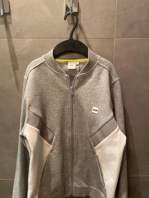 Grå zip-kofta från Hugo Boss - Snygg grå kofta från Hugo Boss med hel dragkedja och coola vita paneler på ärmarna. Diskret BOSS-logga på bröstet och präglad logga längs ryggen och ärmarna. Mjuk bomullsmix och långärmad modell, perfekt för chill eller streetwear.