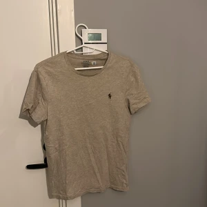 Beige t-shirt från Polo Ralph Lauren - Snygg beige t-shirt från Polo Ralph Lauren med klassisk passform och liten broderad logga på bröstet. T-shirten har rund halsringning och är tillverkad i mjuk bomull, perfekt för en clean och stilren look.