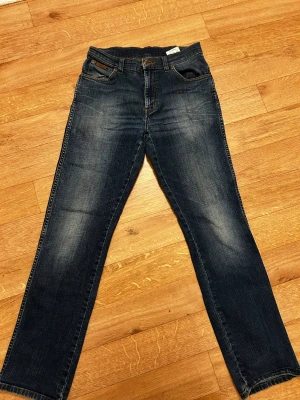 Wrangler blå jeans straight fit - Snygga blå jeans från Wrangler med klassisk straight fit och tydliga sömmar på bakfickorna. Jeansen har fem fickor, bälteshällor och en mörk tvätt med lätt slitning. Materialet är robust denim som ger en cool och tidlös look.