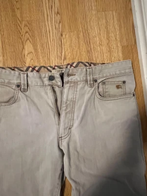 Beige Burberry jeansbyxor, storlek 36 - Snygga beige jeansbyxor från Burberry med klassiskt rutigt foder i midjan och detaljer i rött. Byxorna har fem fickor, dragkedja och knappstängning. Tillverkade i 100% bomull för skön känsla och stilren look. Nypris (5400)