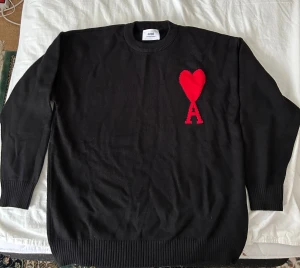 Svart stickad tröja från Ami Paris - Svart stickad tröja från Ami Paris med rund hals och ribbade muddar. På bröstet finns ett stort rött hjärta med bokstaven A, vilket ger en unik och trendig look. Perfekt för dig som gillar stilrena detaljer och vill sticka ut lite extra. Storlek L. Helt ny. 