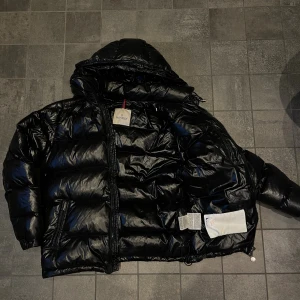 Svart Moncler pufferjacka med huva - Svart pufferjacka från Moncler med glansig finish, huva och dragkedja. Jackan har Moncler-logga på ärmen, praktiska fickor med knappar och quiltad design. Perfekt för kalla dagar och har en riktigt clean look som sticker ut.