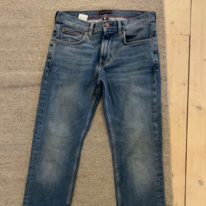 Blå jeans från Tommy Hilfiger - Snygga blå jeans från Tommy Hilfiger med klassisk femficksdesign, bruna detaljer och logga på bakfickan. Nästan inte använt och därför i väldigt bra skick. Jeansen har normal passform och raka ben, tillverkade i slitstarkt bomullsjeans med snygg tvätt och röda sömdetaljer.
