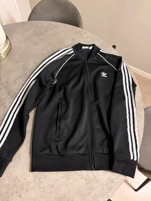 Svart Adidas track jacket L - Svart Adidas track jacket med klassiska vita ränder längs ärmarna och Adidas-logga på bröstet. 