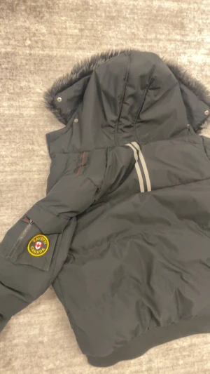 Svart pufferjacka från Zavetti Canada - Svart pufferjacka från Zavetti Canada med stor huva och snygg fuskpäls runt kanten. Jackan har broderad logga på ärmen, två dragkedjefickor fram och coola detaljer med röda sömmar. Perfekt för kalla dagar och har en riktigt schysst streetstil.