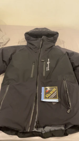 Svart Gore-Tex jacka Sail Racing - Svart jacka från Sail Racing med Gore-Tex material, perfekt för kalla och blöta dagar. Jackan har hög krage, flera dragkedjeförsedda fickor och snygg logga på bröstet. Funktionell design med justerbar huva och robusta detaljer. Grym för dig som gillar stil och funktion.