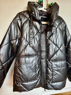 Mörkgrå quiltad pufferjacka från Mohito - Snygg mörkgrå pufferjacka från mohito med glansig finish och quiltat mönster. Jackan har huva, stora fickor framtill och dragkedja. Lite stor för mig så säljer den nästan oanvänd. Oversized passform (bild 3 för bäst bild på färg)