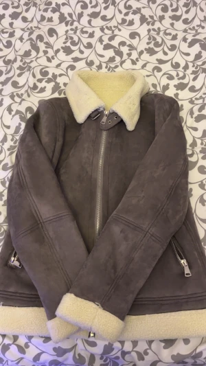 Grå teddyjacka med dragkedja - Säljer en grå teddyjacka med fluffig krage och muddar i offwhite. Jackan har två sidofickor med dragkedja och en snygg metallzip framtill. Materialet är mockaliknande med mjukt teddyfoder som syns vid krage och ärmslut. Perfekt för kalla dagar. Kan gå ner i pris vid snabb affär 