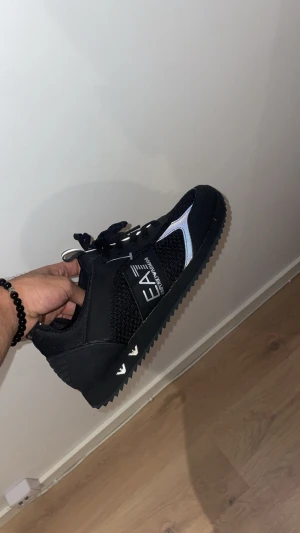 Svarta EA7 Emporio Armani sneakers - Snygga svarta EA7 Emporio Armani sneakers med silvriga detaljer och reflekterande inslag. Ovandelen är i mesh och syntet, med snörning och platt sula med grovt mönster. Logga på sidan och innersulan. Perfekt för dig som gillar stilrena och sportiga skor.