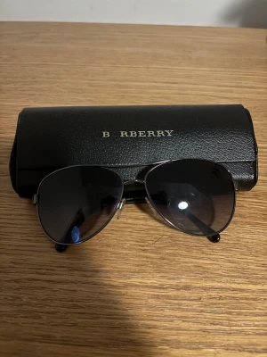Burberry solglasögon - Snygga aviator solglasögon från Burberry med klassisk randig detalj på skalmarna i silver, grått och rött. Glasen är mörktonade och bågarna är i metall med svarta ändar. Kommer med original svart fodral med Burberry-logga.