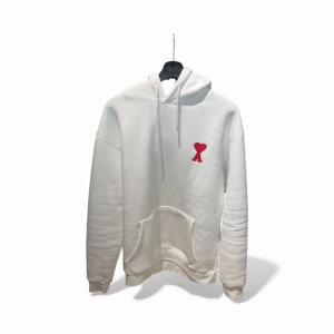 Vit hoodie från Ami med rött hjärta - Vit hoodie från Ami Alexandre Mattiussi i storlek XXL. Tröjan har en röd hjärtlogga med bokstaven A på bröstet, snörning i huvan och en stor magficka. Tillverkad i mjuk bomull med fleece på insidan för extra komfort. Perfekt för chill dagar.