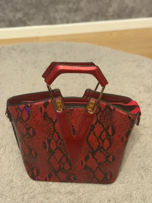 Röd handväska med ormmönster - Snygg röd handväska med svart ormmönster och glansig finish. Väskan har geometriska handtag i rött med guldfärgade detaljer, dragkedja framtill och rymligt fack. Materialet är syntet och designen är riktigt iögonfallande.