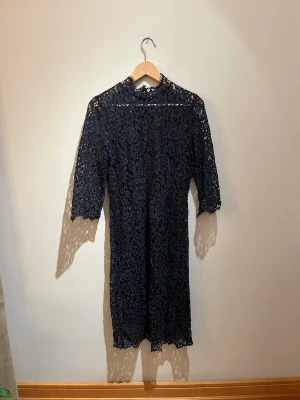 Mörkblå spetsklänning Soaked in Luxury - Supersnygg mörkblå spetsklänning från Soaked in Luxury med blommigt mönster, hög hals och trekvartsärm. Klänningen är fodrad och har en rak passform som slutar strax ovanför knäna. Perfekt för dig som vill sticka ut med elegans.