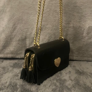 Svart axelväska med hjärtdetalj - Stilren svart axelväska i skinnimitation med guldfärgad kedja och hjärtformad låsdetalj framtill. Väskan har två dragkedjor med tofsar på sidan och en elegant, kompakt form. Perfekt skick 