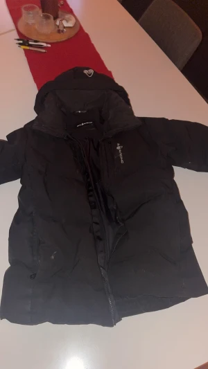 Svart dunjacka Sail Antarctica Expedition - Svart dunjacka från Sail Antarctica Expedition med huva och dragkedja. Jackan är fodrad med Allied Feather Down 500 fill power och har en stilren look med diskret logga på bröstet. Perfekt för kalla vinterdagar och riktigt skön passform. En av dragkedjorna e sönder på bröstfickan.