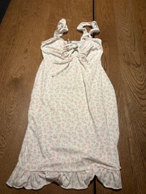 Blommig klänning från Nelly - Supersöt vit klänning med ljusrosa blommor från Nelly. Den har volangkant nertill, rynkade axelband och snörning vid bysten. Perfekt för sommarens alla tillfällen! Mjuk bomullskänsla och riktigt gullig passform.