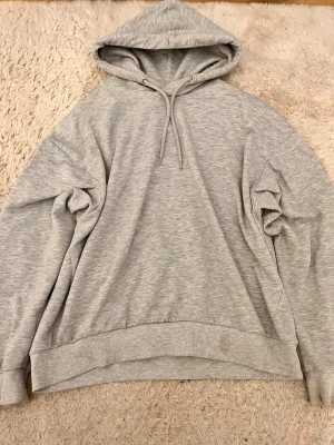 Grå hoodie med huva och snörning - Säljer en enkel och stilren grå hoodie med huva och snörning. Tröjan har en loose passform och är tillverkad i mjuk bomullsmix. Perfekt för chill dagar och passar till det mesta. Ribbad mudd vid ärmslut och nederkant för extra komfort.