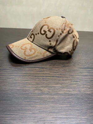 Gucci beige keps med GG-mönster - Snygg beige keps från Gucci med det klassiska GG-mönstret i brunt och ljusbrunt. Kepsen har en böjd skärm med brun kant och är tillverkad i bomull och polyester. Perfekt för dig som vill ha en lyxig och trendig accessoar med tydlig logga.