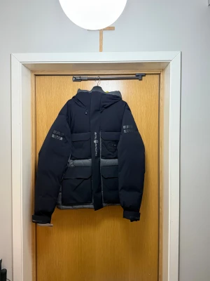 Svart Sail Racing dunjacka med huva - Säljer en svart och grå dunjacka från Sail Racing med huva och feta loggor på ärmarna. Jackan har stora fickor framtill, dragkedja och är fodrad med 700 fill power dun från Allied Feather + Down. Perfekt för kalla vinterdagar och har en clean, sportig look.