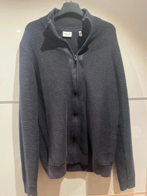 Mörkblå ribbad kofta från Solid - Säljer en mörkblå kofta från Solid i storlek M. Tröjan har ett ribbat, randigt mönster och dragkedja framtill. Hög krage och långa ärmar ger en chill och stilren stil.