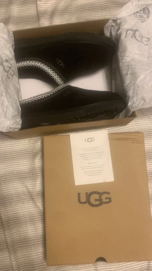 Svarta UGG boots i mocka - Svarta UGG boots i mjuk mocka med fluffigt foder och dekorativt mönster runt öppningen. Skorna har rund tå, platt sula och UGG-logga på sidan. Perfekta för kalla dagar när du vill ha både stil och komfort.