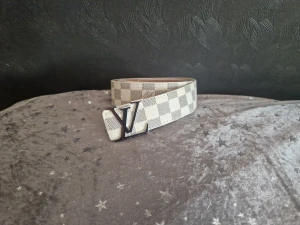 Louis Vuitton rutig bälte i skinn - Säljer ett snyggt Louis Vuitton bälte med klassiskt rutigt mönster i beige och vitt. Bältet har en stor LV-logga i silver som spänne och är tillverkat i skinn. Perfekt accessoar för att lyfta din outfit.