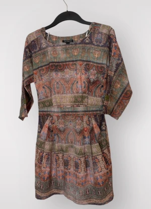Mönstrad kortklänning från MNG - Unik kortklänning från MNG med färgstarkt paisleymönster i jordnära toner av orange, grön, blå och beige. Klänningen har trekvartsärm, rund hals och lätt rynkad midja för en avslappnad men snygg siluett. Tillverkad i mjuk bomull, perfekt för vår och sommar.