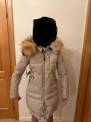 Cedrico Vinterjacka med päls - Snygg beige dunjacka med fluffig päls runt luvan och dragkedja framtill. Jackan har långa ärmar, flera fickor med dragkedja och en figurnära passform med snörning i midjan. Perfekt för kalla vinterdagar och ger en cool look. Knappt använd dessutom:)