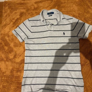 Grå randig pikétröja från Polo Ralph Lauren - Snygg grå pikétröja från Polo Ralph Lauren i storlek S. Tröjan har mörkgrå horisontella ränder, klassisk krage och två knappar vid halsen. Ikoniska broderade loggan på bröstet. Tillverkad i mjuk bomull och har korta ärmar. Perfekt för en chill och stilren look.