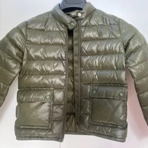 Olivgrön dunjacka från Polo Ralph Lauren - Säljer en olivgrön dunjacka från Polo Ralph Lauren. Jackan har quiltad design, två stora fickor framtill med knappar, hög krage med tryckknapp och dragkedja. Perfekt för kyliga dagar och riktigt snygg till jeans eller hoodie. Storleken är en XL men passar ungefär S-M med korta armar.