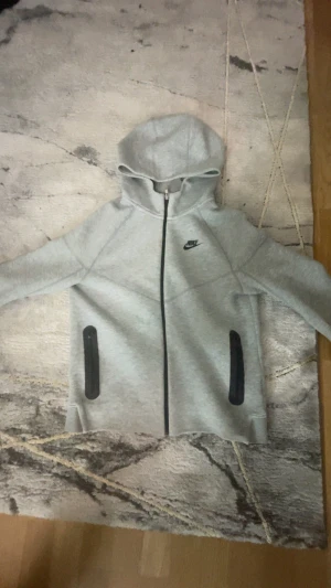 Grå hoodie från Nike med dragkedja - Säljer en grå hoodie från Nike med hel dragkedja och svart logga på bröstet. Tröjan har huva, två svarta fickor med dragkedja och långärmad design. Perfekt för chill eller träning, riktigt skön och stilren look.