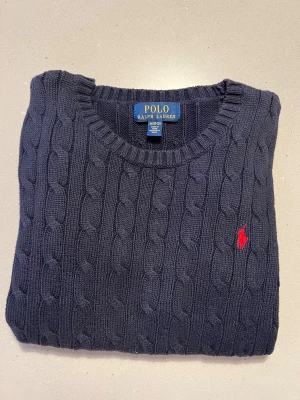 Mörkblå stickad tröja Polo Ralph Lauren - Snygg mörkblå stickad tröja från Polo Ralph Lauren med klassiskt kabelstickat mönster och röd broderad logga på bröstet. Rund halsringning och långa ärmar med ribbade muddar. Perfekt för dig som gillar stilrena och tidlösa plagg.