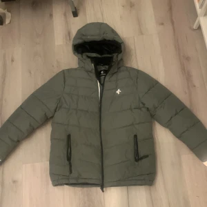 Grå dunjacka från Cross Sportswear - Säljer en grå dunjacka från Cross Sportswear. Jackan har en huva och dragkedja framtill samt två praktiska fickor med dragkedjor. Perfekt för kyliga dagar.