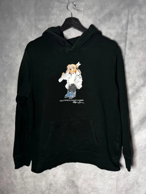 Svart Polo Bear hoodie från Ralph Lauren - Svart hoodie från Ralph Lauren med Polo Bear-tryck på bröstet. Tröjan har huva, känguruficka och långa ärmar. Materialet är mjuk bomull och passformen är normal. Perfekt för dig som gillar streetwear och vill ha en chill look.Storlek 18-20 sitter som S. Inga fläckar eller hål men använd en del