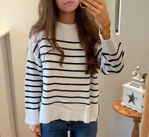 Randig vit stickad tröja från 157 - Supermysig vit stickad tröja med svarta horisontella ränder från 157. Tröjan har rund hals, långa ärmar och breda ribbade muddar vid ärmslut och nederkant. Perfekt för lager-på-lager och chill vardagsstil.