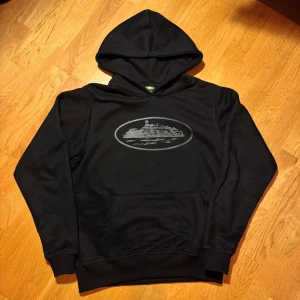 Svart hoodie med tryck, Small - Svart hoodie i storlek Small med ett grått tryck av en byggnad på bröstet. Tröjan har huva, långärmade ärmar och en klassisk magficka framtill. Materialet känns mjukt och är troligen bomull eller polyester. Perfekt för chill dagar och streetstyle.