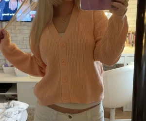 Persikofärgad stickad kofta med knappar - Mysig persikofärgad stickad kofta med v-ringning och stora matchande knappar framtill. Koftan har en loose passform och långa ärmar, perfekt att slänga på sig när det är lite kyligt. Ribbad struktur ger en skön och trendig look. Storlek 164 men uppskattas till XS-S.