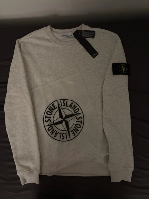 Stone Island ljusgrå sweatshirt M - Ljusgrå sweatshirt från Stone Island i storlek M. Klassisk rund hals och ribbade muddar. Ikonisk Stone Island patch på vänster ärm och stor logga på framsidan. Mjuk bomullskvalitet som är skön att bära. Perfekt för dig som gillar streetwear och snygga detaljer. PRISET KAN DISKUTERAS 