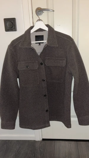 Brun overshirt från 157, storlek S - Hej! Säljer en stilren brun overshirt från 157 i storlek S. Jackan har klassisk krage, två bröstfickor med knappar och knäppning framtill. Materialet är mjukt och känns som ull, perfekt för lager-på-lager. Passformen är normal och ärmarna är långa.