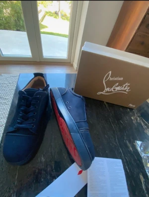 Mörkblå sneakers från Christian Louboutin - Snygga mörkblå sneakers från Christian Louboutin med klassisk röd sula och rund tå. Skorna har snörning och är tillverkade i mocka med detaljer i skinn. Insidan är beige och de har en platt sula. Perfekt för dig som gillar exklusiva märken och stilren design.