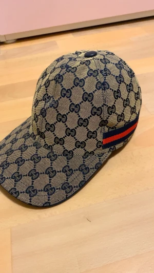 Gucci keps - Snygg beige keps från Gucci med klassiskt GG-mönster i mörkblått och en röd-blå rand på sidan. Justerbar baktill med metallspänne. Perfekt för dig som vill ha en lyxig och trendig accessoar till din outfit.