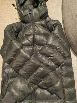 Mörkgrön dunjacka från Peak Performance - Snygg mörkgrön dunjacka från Peak Performance med huva och dragkedja framtill. Jackan har ett quiltat mönster och ribbade muddar vid ärmslut. Perfekt för kalla dagar och har en clean, sportig look med diskret logga på bröstet. Nästan aldrig andvänd.