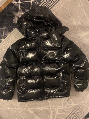 Svart pufferjacka från Trapstar - Vi kan diskutera pris 1500 Säljer en svart, glansig pufferjacka från Trapstar med huva och fet logga broderad på ryggen. Jackan har dragkedja framtill, två stora fickor och coolt emblem på bröstet. Perfekt för dig som vill sticka ut med streetstyle i vinter. Har använt 0 gångee