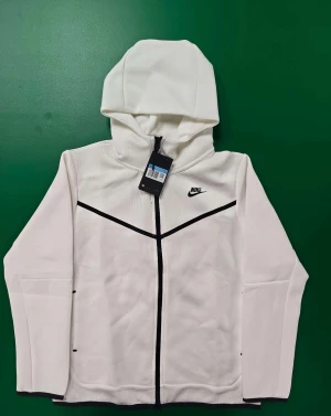Vit Nike tech - Nike tech i vit med svarta detaljer och dragkedja framtill. Jackan har en sportig look med huva, lång ärm och svarta linjer som ger en snygg kontrast. Perfekt för dig som gillar stilren och modern design.