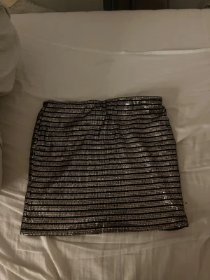 Guldig paljettkjol från Amisu - Säljer en kort kjol från Amisu med glittriga guldpaljetter och svarta ränder. Kjolen har en rak passform och är fodrad med svart tyg på insidan. Perfekt för dig som vill sticka ut med lite extra bling och coola detaljer.