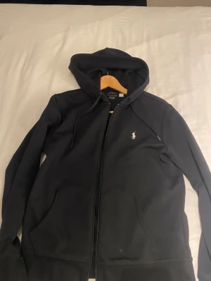 Svart hoodie från Polo Ralph Lauren - Svart hoodie från Polo Ralph Lauren med klassisk logga broderad på bröstet. Tröjan har huva med snörning, dragkedja framtill och två fickor. Tillverkad i mjuk bomull, perfekt för chill dagar. Snygg och enkel design som funkar till allt.