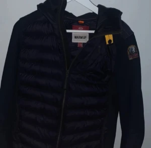 Svart pufferjacka Parajumpers - Svart pufferjacka från Parajumpers med quiltad framsida och rygg, samt släta ärmar. Jackan har en gul detalj vid kragen, dragkedja framtill och en patch på ärmen. Perfekt för dig som vill ha en snygg och funktionell jacka med cool design.