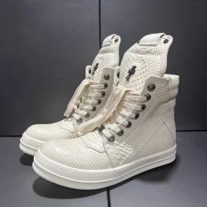 Vit high-top boots med reptilmönster - Unika high-top boots i vit färg med reptilmönstrad yta och metallkorsdetaljer på plösen. Skorna har snörning, dragkedja på sidan och robust platt sula. Materialet ser ut som präglat skinn och ger en lyxig vibe. Perfekt för dig som vill sticka ut med din stil.