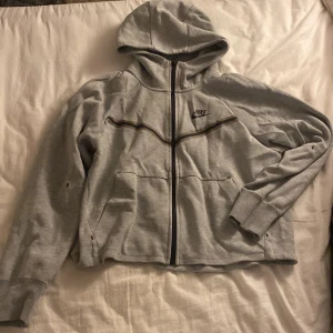 Grå zip-hoodie från Nike - Grå hoodie från Nike med hel dragkedja och huva. Jackan har svarta detaljer längs bröstet, två fickor framtill och ribbade muddar. Perfekt för chill dagar eller när du vill ha en sportig look. Helt oanvänd 